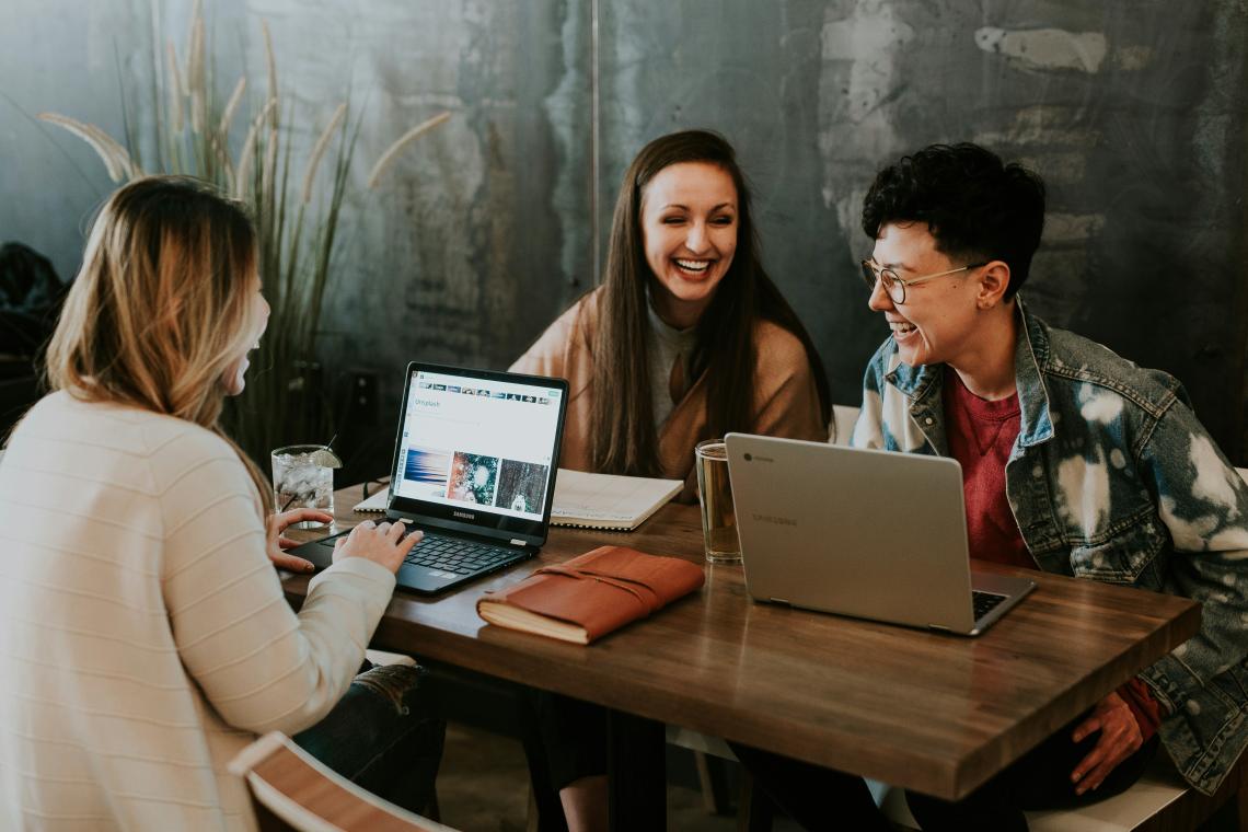 Three people sit at a wooden table with laptops, smiling and engaging in a lively discussion.