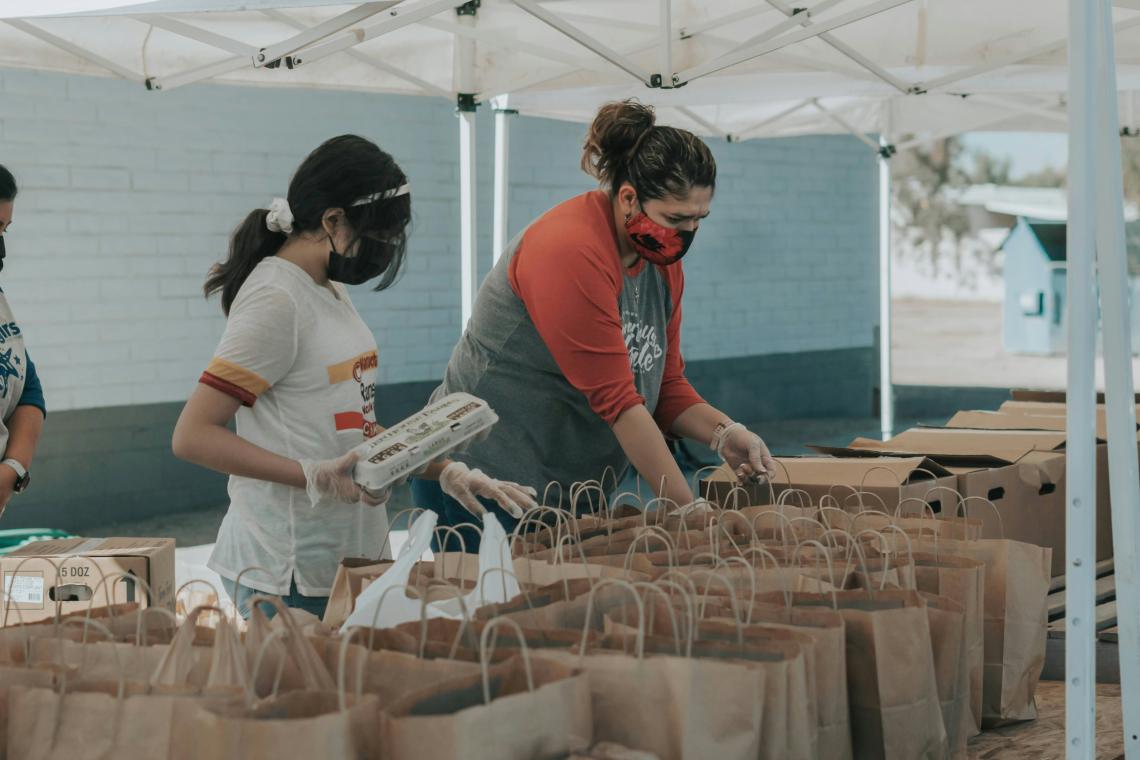 Two volunteers in masks and gloves prepare food packages under a tent.