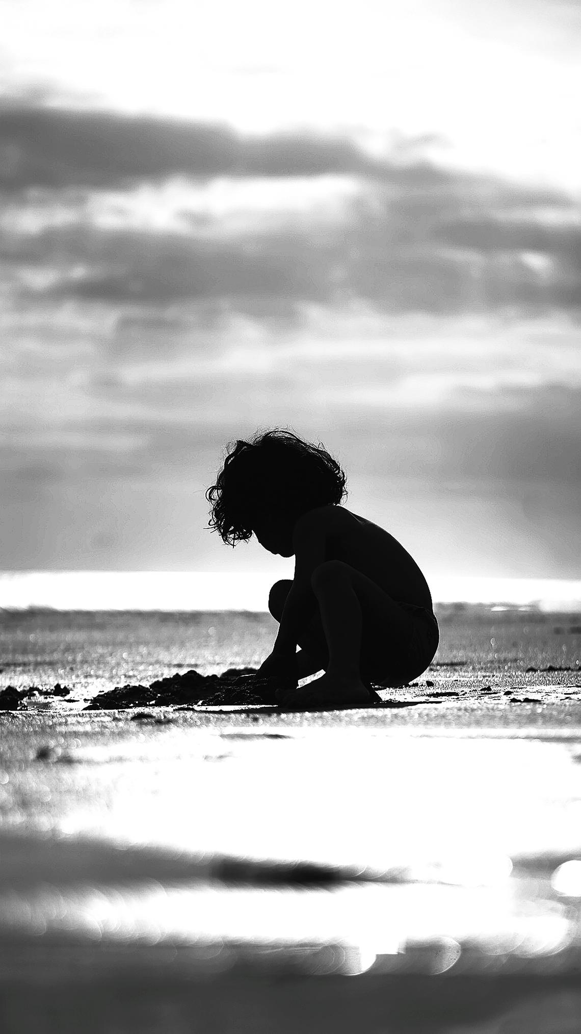 Black and white picture with a silhouette of a child crouching on a beach, playing in the sand.