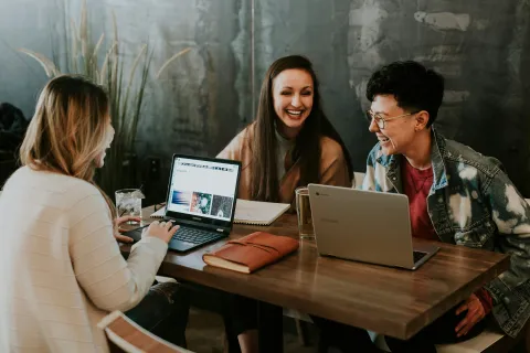 Three people sit at a wooden table with laptops, smiling and engaging in a lively discussion.