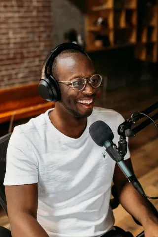 Smiling man with glasses and headphones speaks into a microphone in a cozy, wood-accented room.