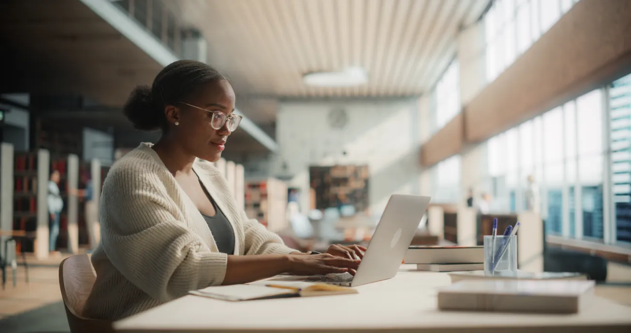 Female Student Engaged in Online Learning at a Modern Library. Young Woman Using Laptop for Research, Surrounded by Books and Academic Environment.