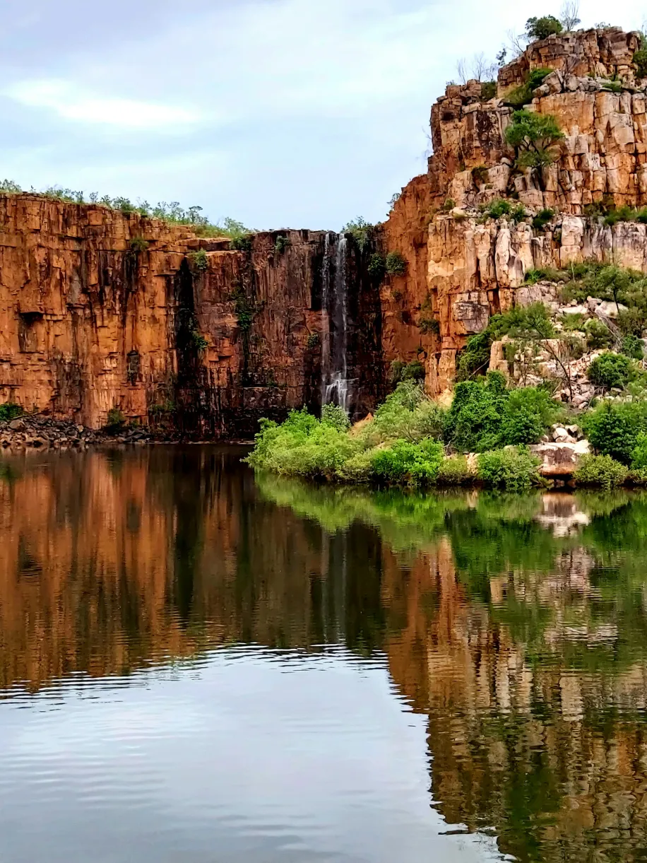 Waterfall in Kimberley, Western Australia, cascades down cliffs into a lake. Greenery surrounds the rocky landscape.